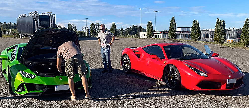 En lysegrønn Lamborghini med åpent bagasjerom foran og en rød Ferrari parkert på en asfaltert plass under en blå himmel.