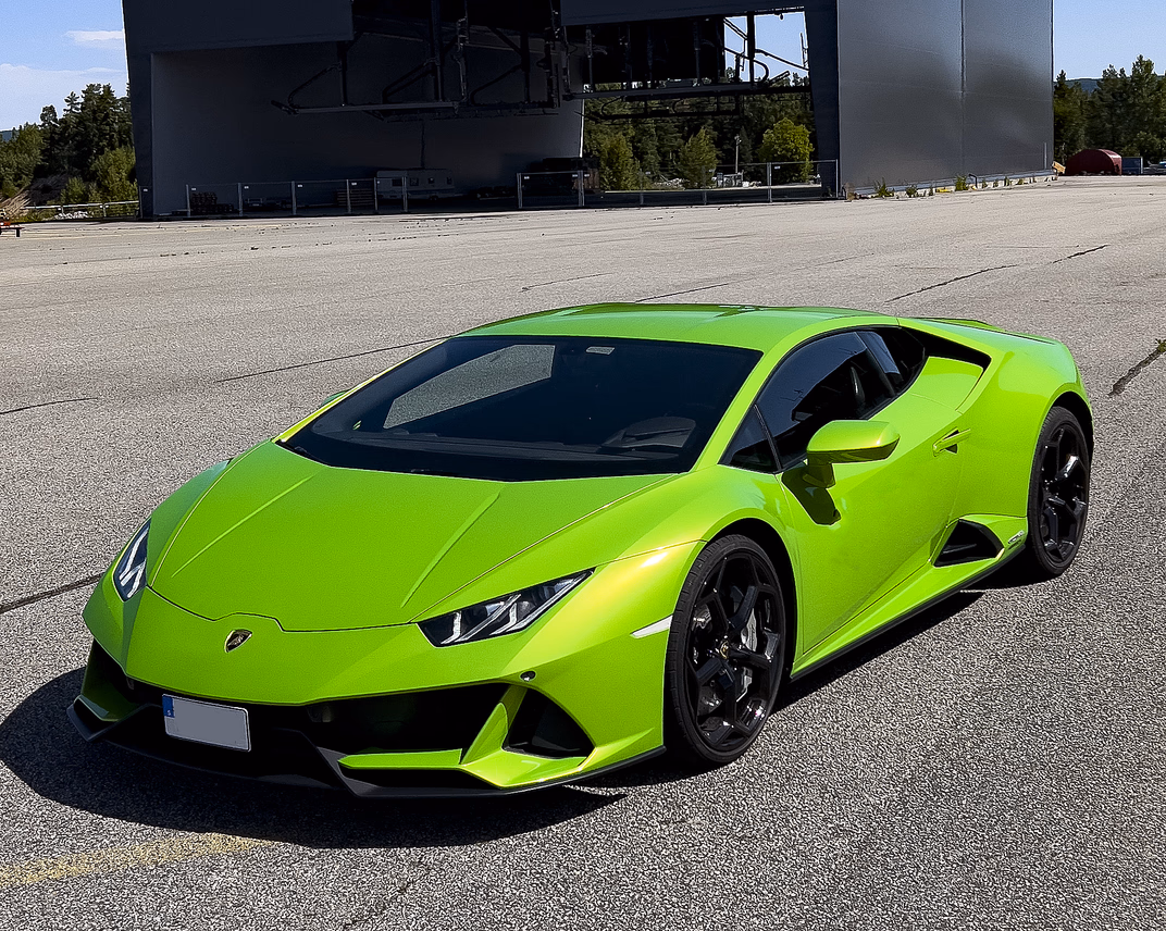 En lys limegrønn Lamborghini sportsbil med skarpe linjer og svarte felger, parkert på en åpen asfaltplass foran en stor grå hangar merket "MG Production AS".