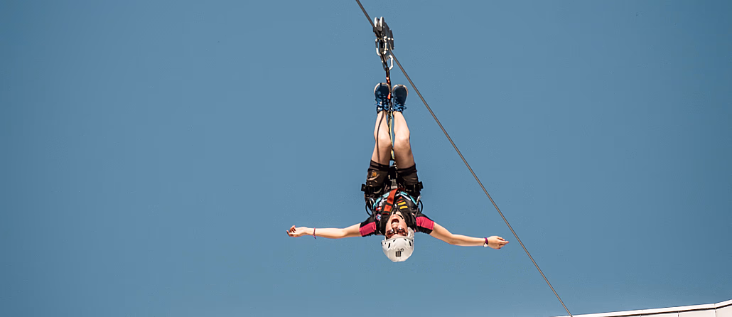 Person i rød skjorte henger opp ned på en zipline med utstrakte armer mot en blå himmel