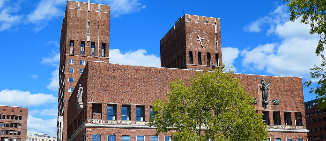 Stor rød mursteinsbygning med to høye tårn, der det høyre tårnet har en stor klokke. Foran er en steinbelagt plass med statuer og grøntarealer under en blå himmel.