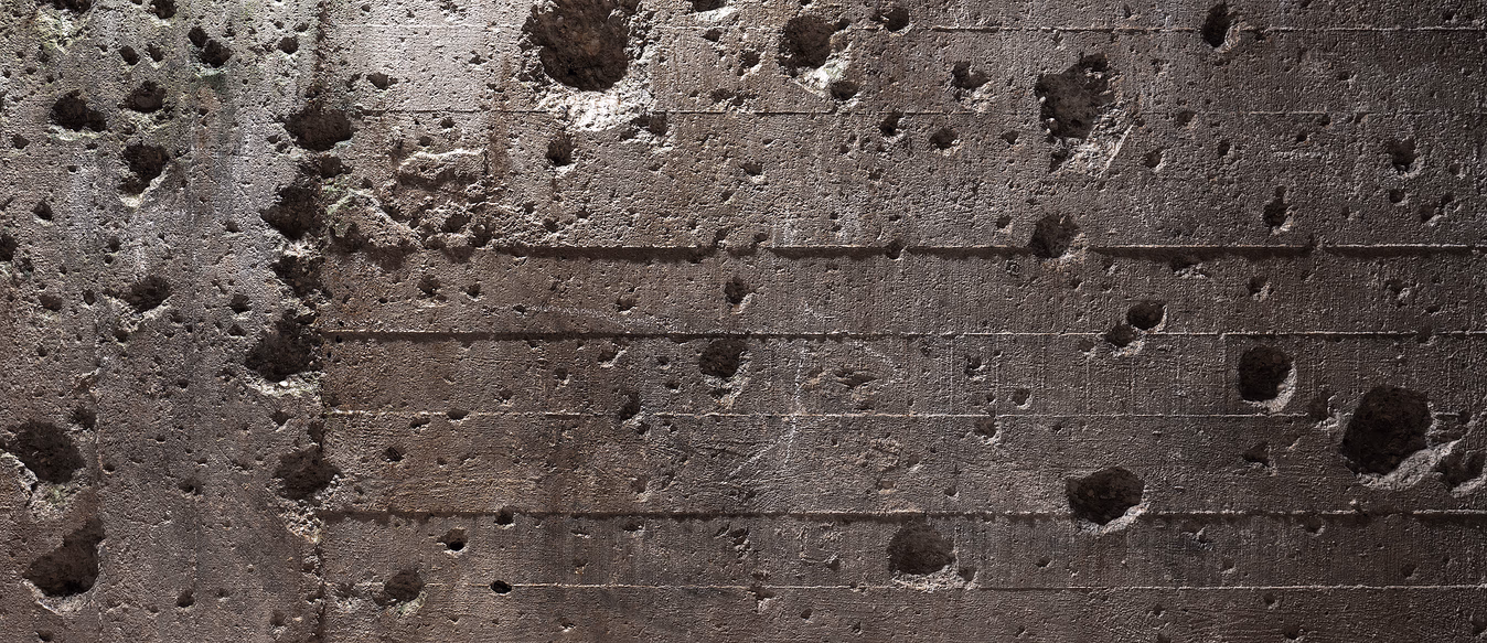 Grov grå betongvegg med små hull, kratre og svake horisontale linjer, med innslag av brune toner og grønne moseflekker.