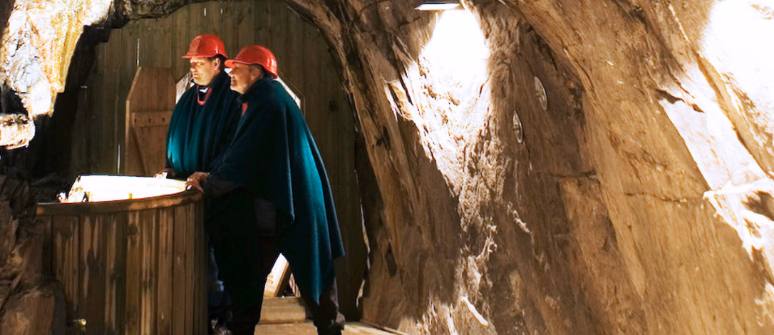 To personer i røde hjelmer og mørkegrønne kapper står ved et tregelender i en opplyst grotte