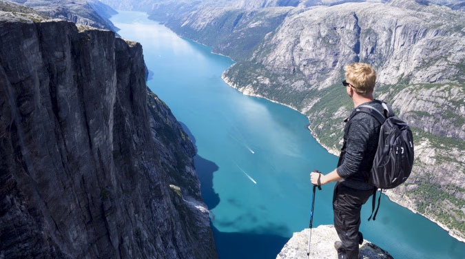 Turgåer med ryggsekk og vandrestaver på en klippe med utsikt over en blå fjord mellom bratte fjell