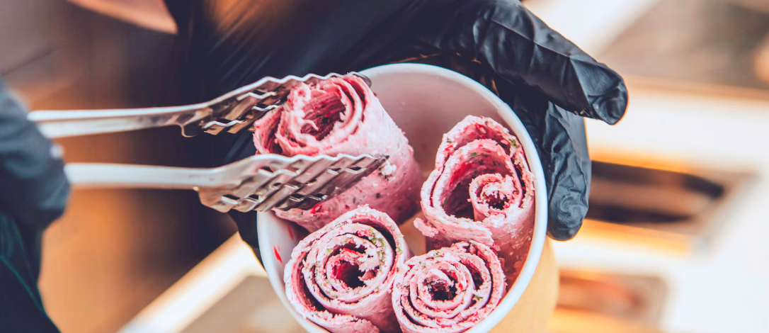 Person med svarte hansker holder et beger med rosa rulleis og bruker en metallklype