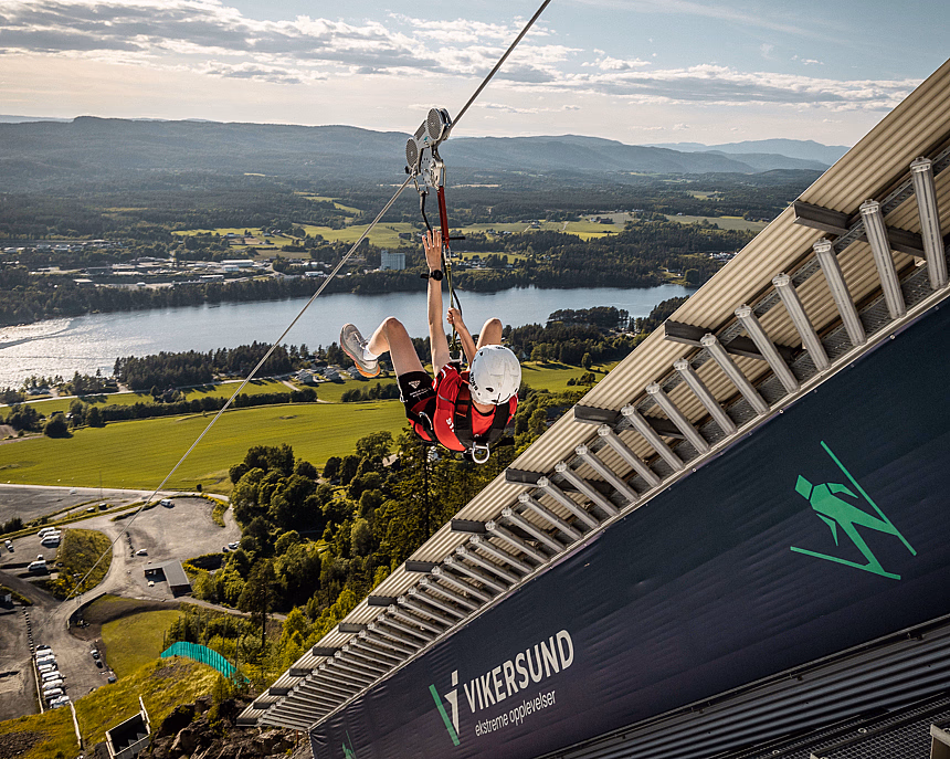 Person i rød skjorte kjører zipline fra tårn merket Vikersund over grønt landskap og vann