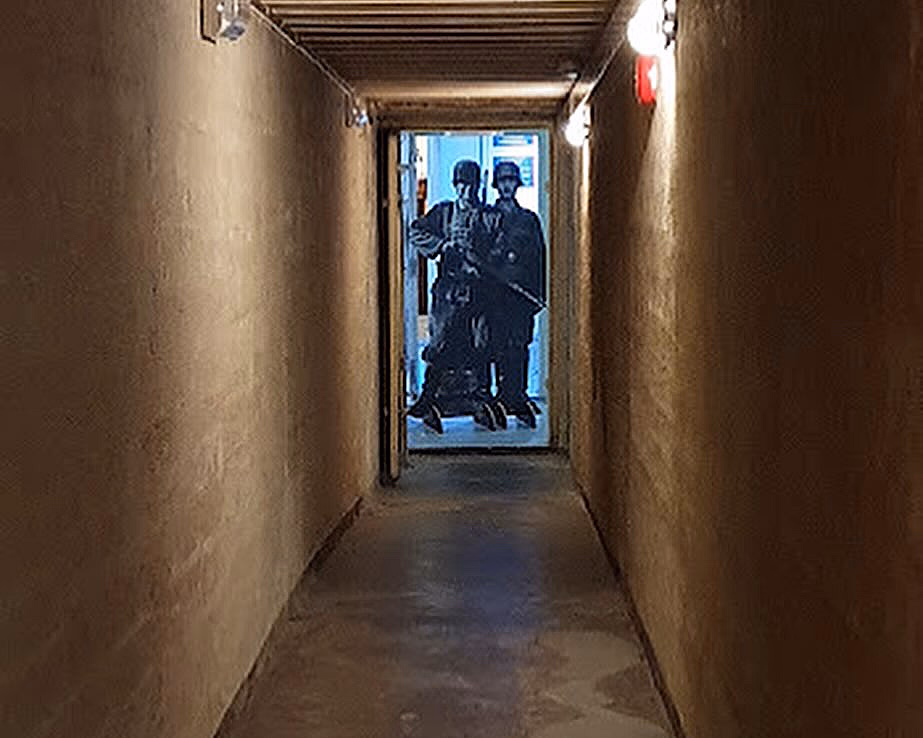 En smal, mørk korridor med grove vegger og betonggulv, som leder til en døråpning med blått lys og figurer av soldater i uniform.