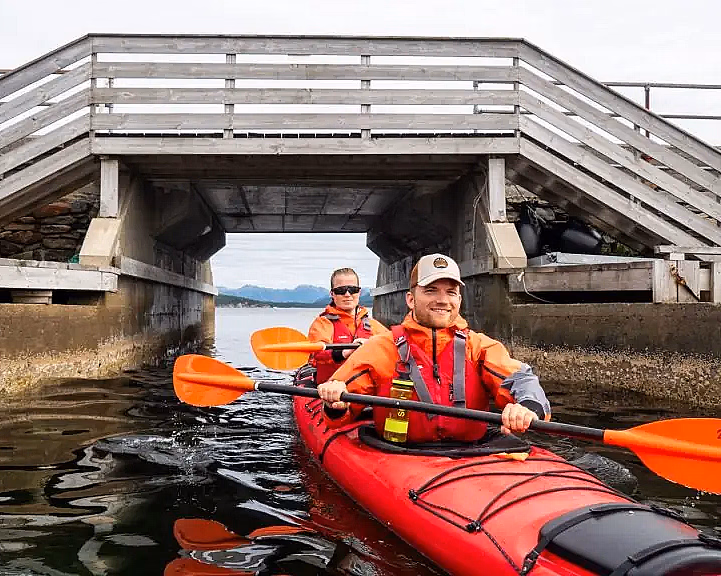 To personer i en rød tandemkajakk padler under en lav trebro i en smal kanal. De har på seg oransje redningsvester og bruker oransje årer, med blå åser og en overskyet himmel i bakgrunnen.
