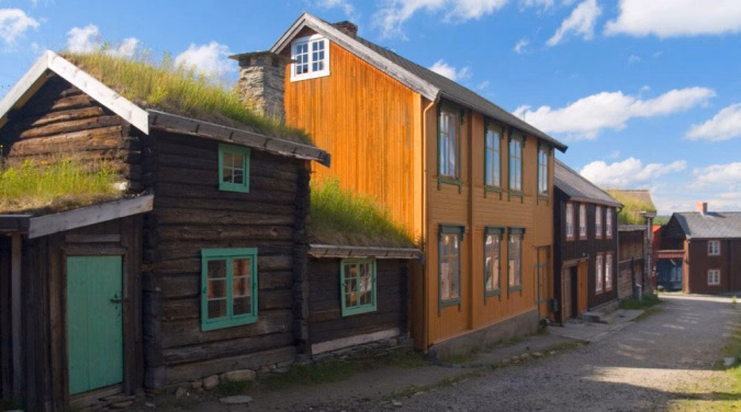 Mørkebrunt trehus med torvtak og grønne vinduer ved siden av en oransje trebygning