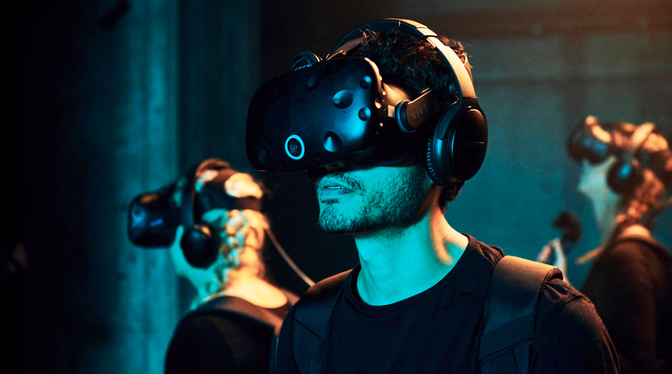 Person med svart VR-headset og hodetelefoner i blågrønt lys med andre i bakgrunnen