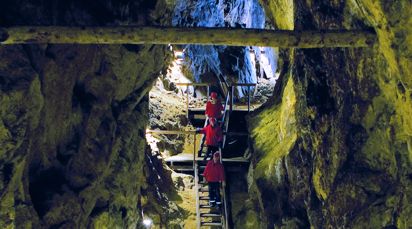 Tre personer i røde jakker og hjelmer går ned en tretrapp i en grotte