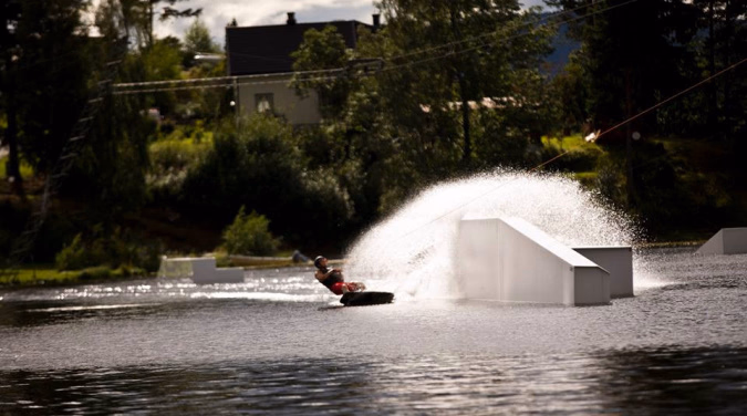 Person på wakeboard på en innsjø ved siden av en hvit rampe med et stort vannsprut