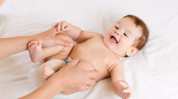 Smilende baby på hvitt teppe som rekker tunge mens voksne hender holder beina