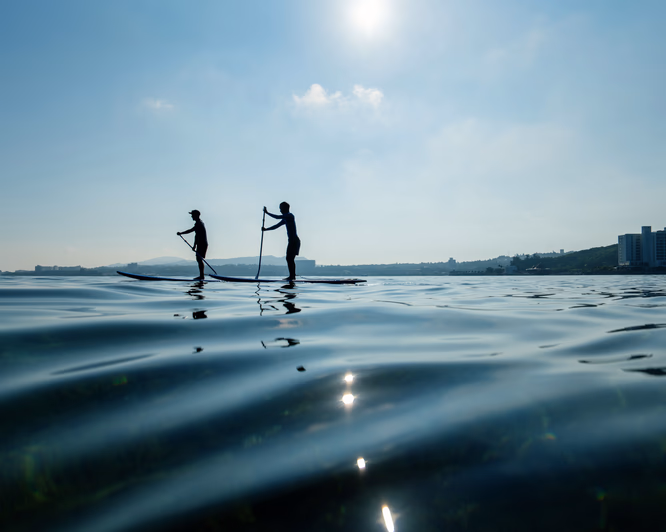 To personer på padlebrett i silhuett på rolig vann med sol og blå himmel