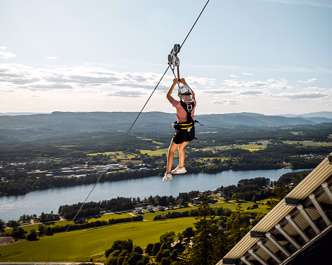 Person i sele og hjelm som kjører zipline over en elv og grønt landskap