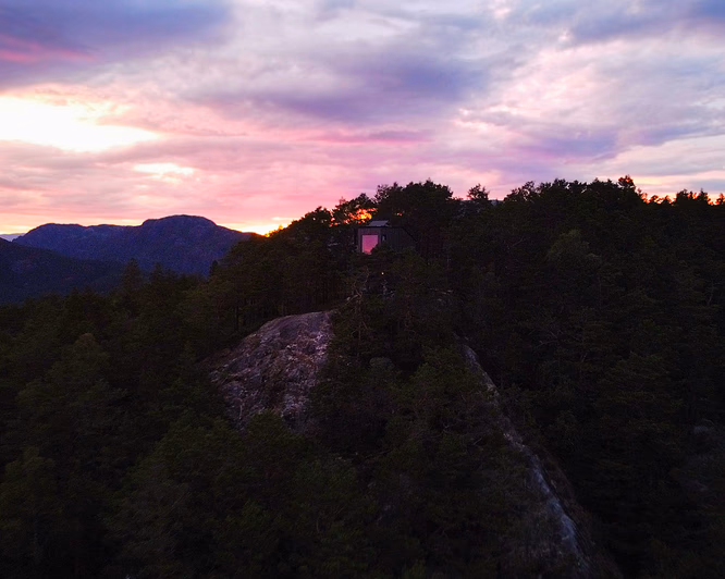 En liten mørk hytte på en steinete åsrygg omgitt av furuskog, med en fargerik solnedgang i lilla, rosa og oransje. Et vindu i hytta lyser opp i rosa, med fjerne fjell i bakgrunnen.