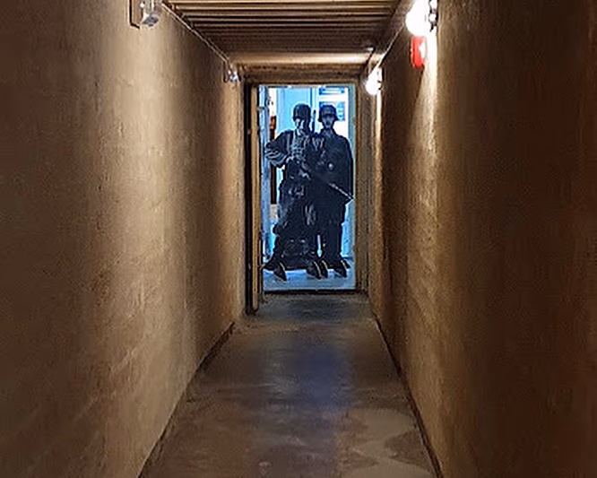En lang, smal og dunkel gang med grove vegger og tretak. I enden er en døråpning opplyst i blått lys, med figurer av soldater i uniform.