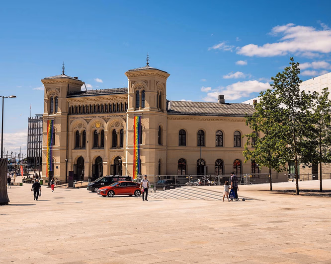 Nobels Fredssenter, en beige steinbygning med to tårn og regnbuebannere, sett fra en stor plass under en blå himmel.