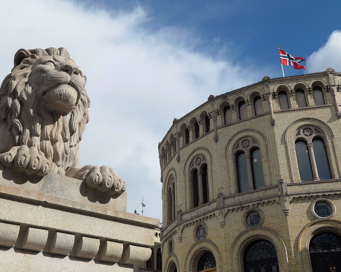 Stor løvestatue i stein foran en rund gul mursteinsbygning med buevinduer og det norske flagget på taket under en blå himmel.