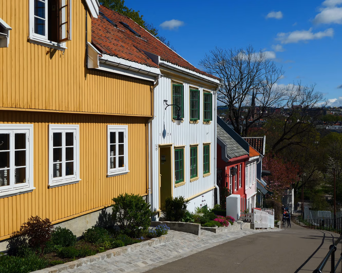 Bratt gate med fargerike trehus i gult, hvitt og rødt, blomsterbed langs veikanten og to syklister på vei ned bakken.