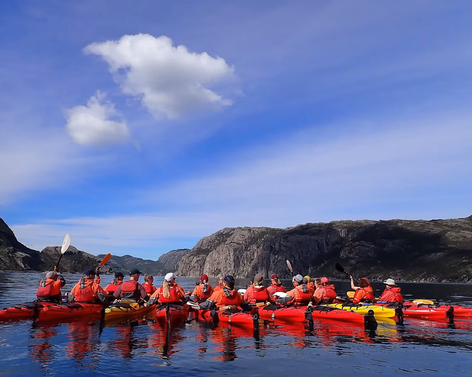 En stor gruppe padlere i røde jakker og fargerike kajakker på en rolig blå fjord, omgitt av bratte klipper og grønne fjellsider.