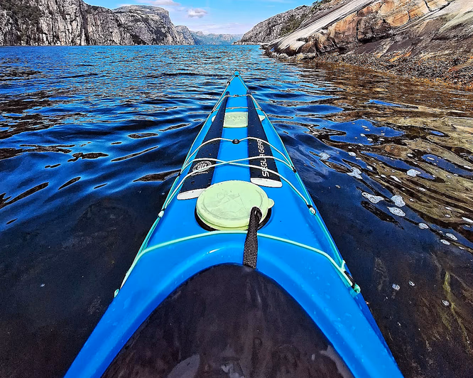 Blå kajakkbaug på mørkt vann i en fjord omgitt av bratte fjellvegger under en blå himmel med lette skyer.