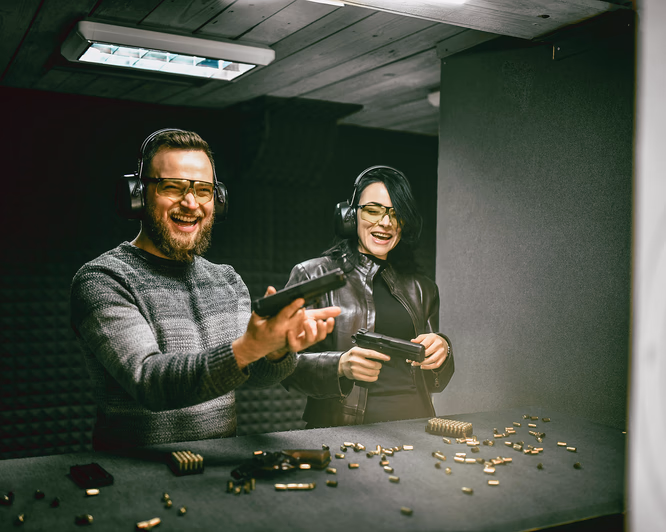 To personer med hørselvern og gule vernebriller holder pistoler på en innendørs skytebane.
