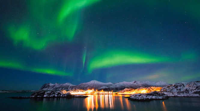 Grønt nordlys over snødekte fjell og en liten by som speiler seg i vannet