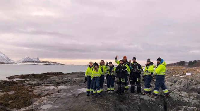 Gruppe i gule jakker på steiner ved havet med snødekte fjell i bakgrunnen