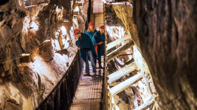 Tre personer med røde hjelmer og kapper går på en trebro i en belyst grotte