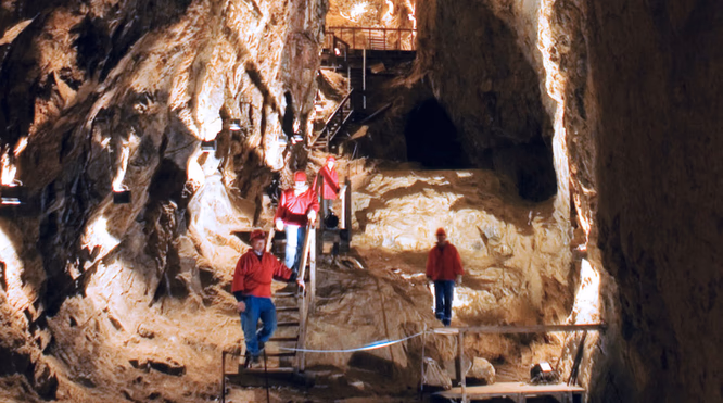 Fire personer med hjelmer og røde jakker går på en tretrapp i en belyst grotte