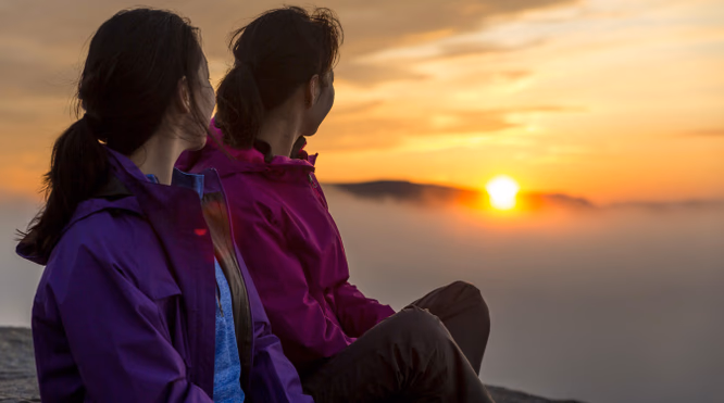 To personer i lilla og rosa jakker sitter på et berg og ser mot en oransje solnedgang