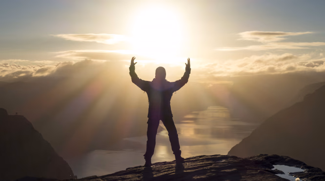 Silhuett av en person med hevede armer på en klippe med utsikt over fjell og vann i solnedgang