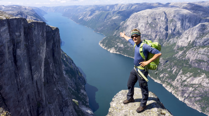 Person med grønn sekk står på en klippe og ser utover en fjord omgitt av bratte fjell