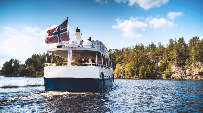 Hvit og blå båt med passasjerer på en innsjø foran skogkledde klipper