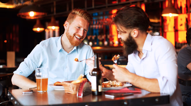To menn som smiler og spiser ved et restaurantbord med mat og drikke